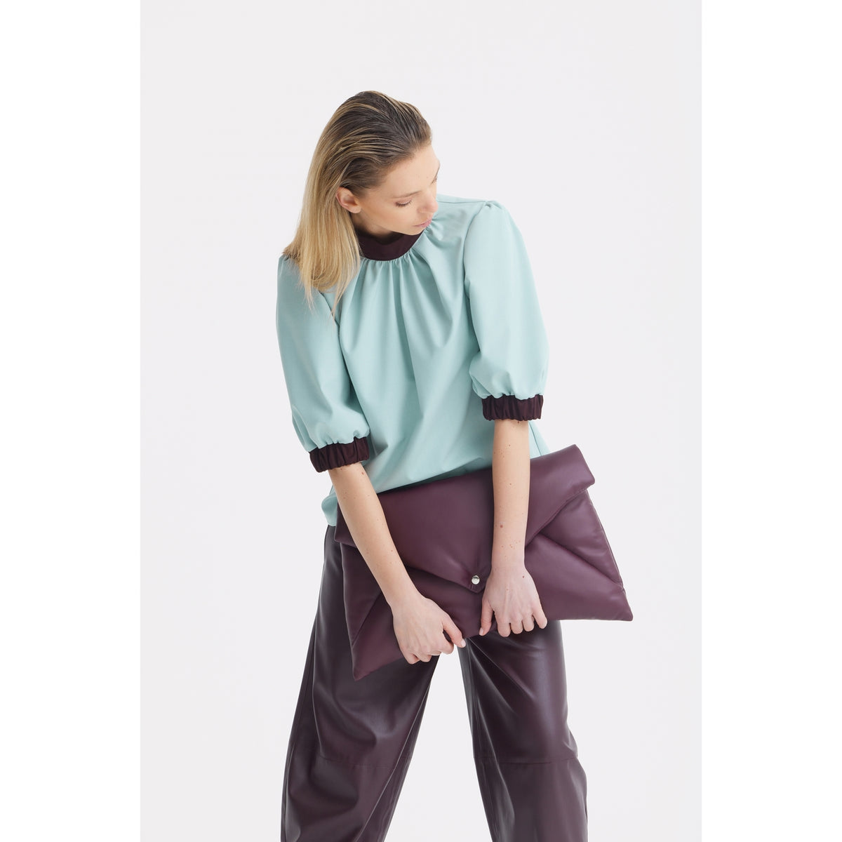 A person with straight blonde hair wears JULIA ALLERT's Textured Short Sleeve Blouse in light blue and loose dark pants, holding a large folded dark purple clutch while looking down against a white background.