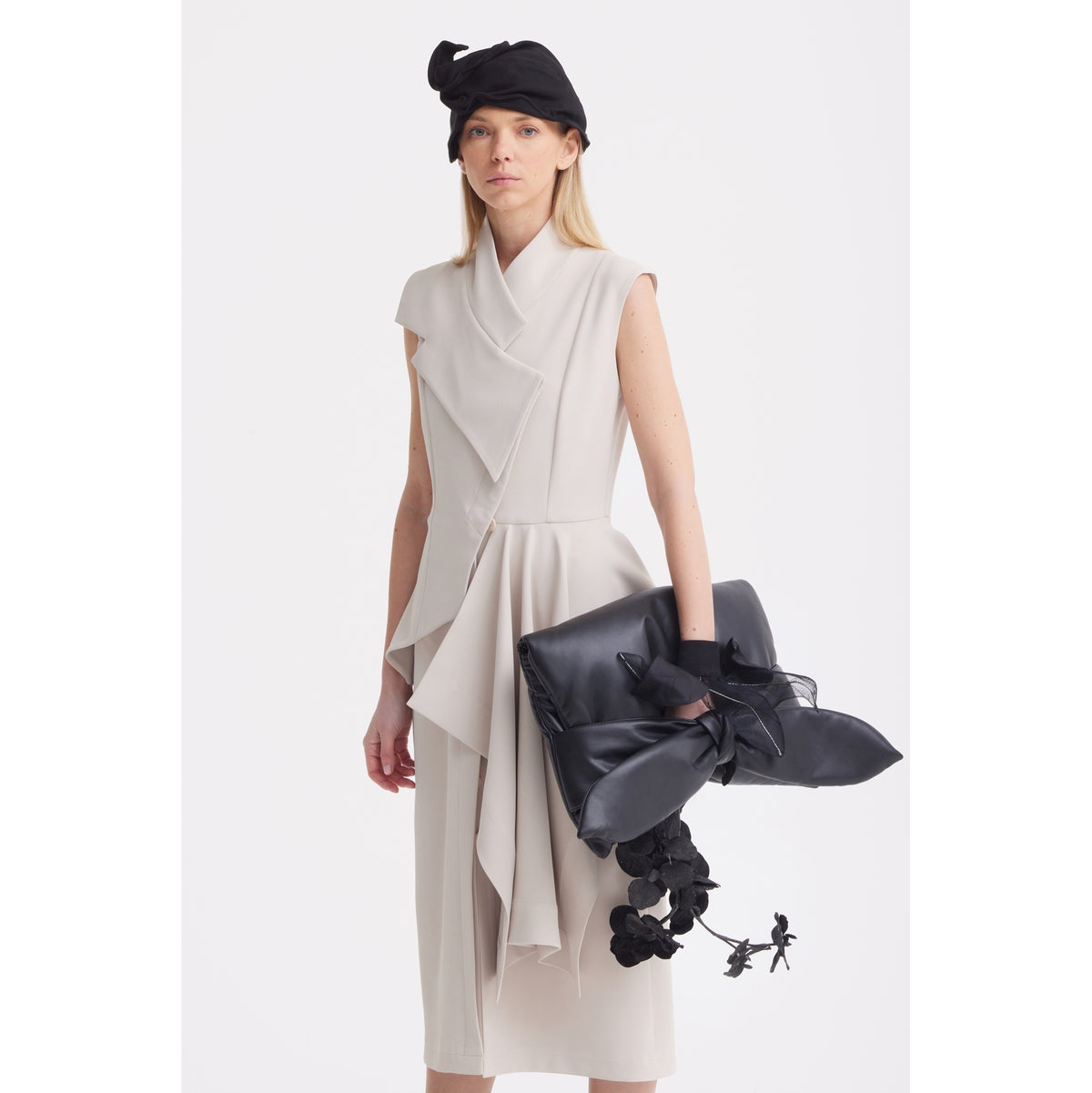 A woman stands against a white background wearing the JULIA ALLERT Architectural Draped Midi Dress in beige, styled sleeveless and button-front. She pairs it with a black headpiece and holds a large black clutch featuring a big bow and floral details.
