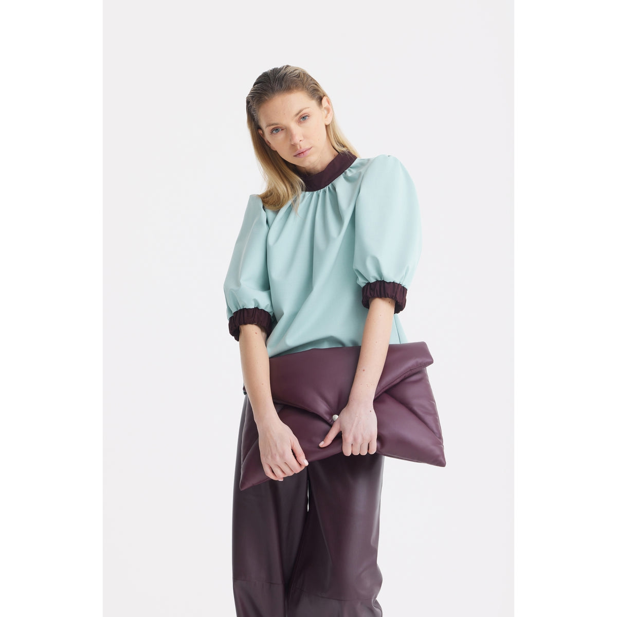A woman with long blonde hair wears a JULIA ALLERT Textured Short Sleeve Blouse in light blue and holds a large, folded dark purple clutch. She stands against a plain white background.