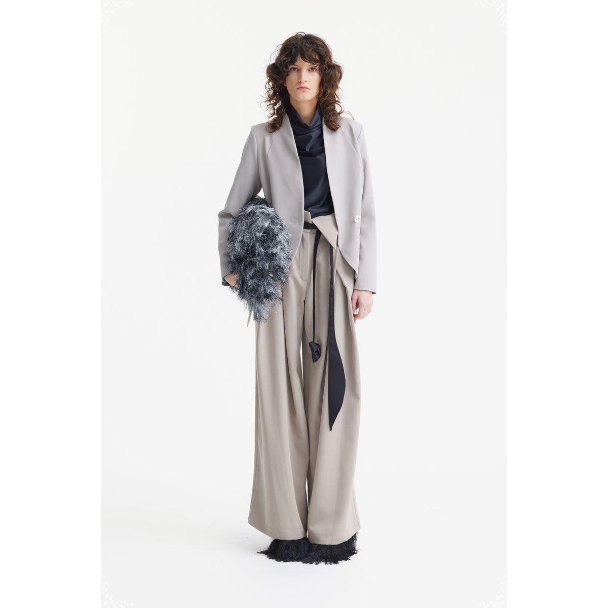 A woman with wavy hair wears JULIA ALLERT's Fashion Forward Wide-Leg Pants &amp; Contrast Belt in beige, styled with a light gray blazer, black top, and textured gray clutch, standing against a plain white background.