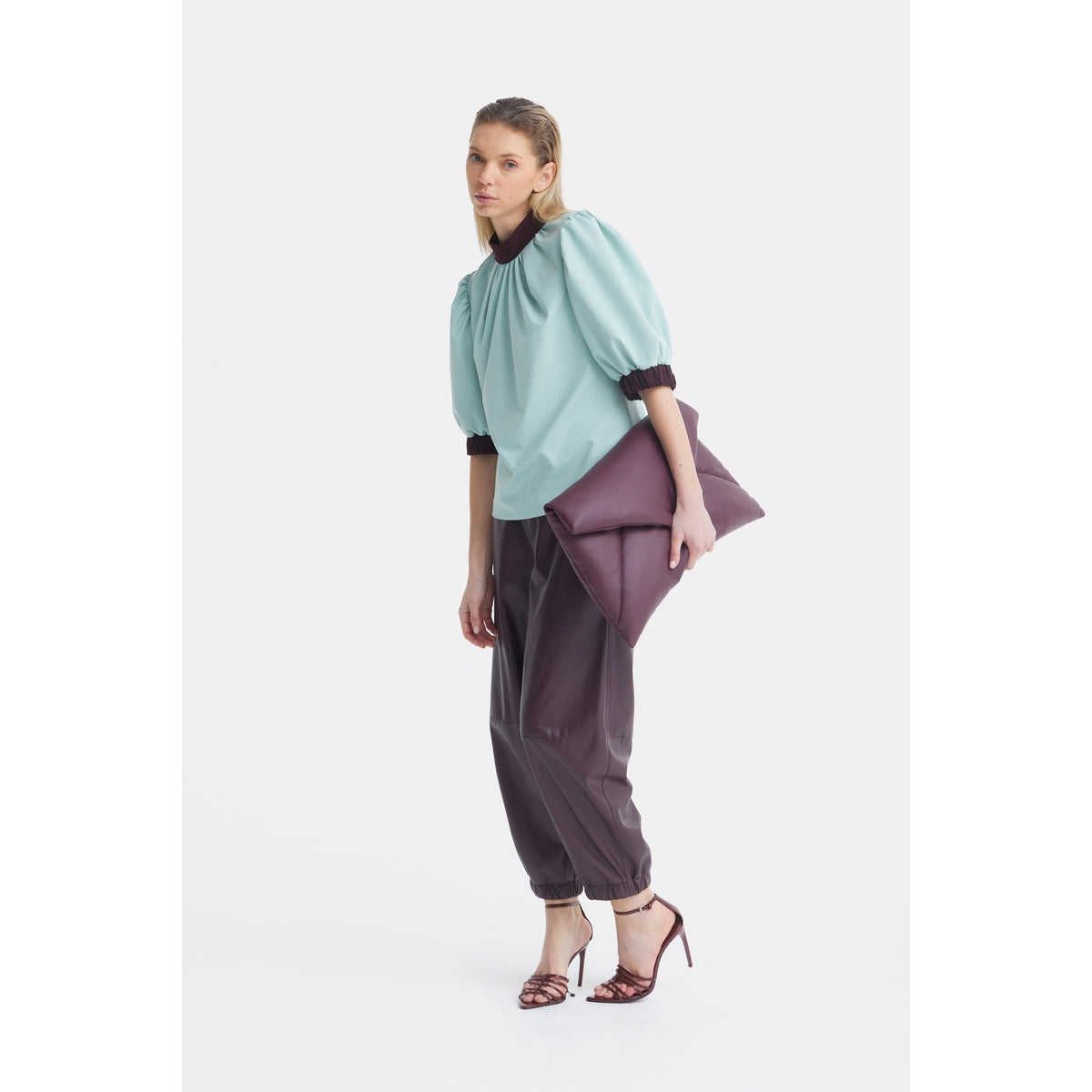 A woman poses against a white background in JULIA ALLERT's Textured Short Sleeve Blouse in light blue, paired with loose dark pants and strappy heels. She carries a large plum clutch, effortlessly combining style with breathable comfort.