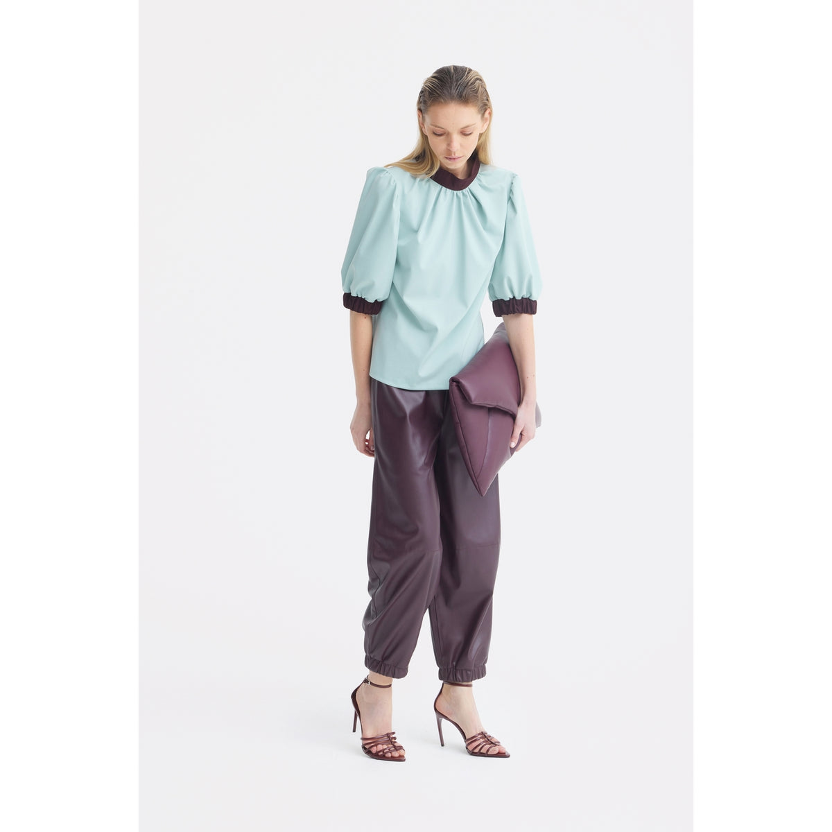 A woman in a light blue Textured Short Sleeve Blouse by JULIA ALLERT, dark burgundy pants, and strappy heels holds a matching clutch, standing against a plain white background and looking downward.