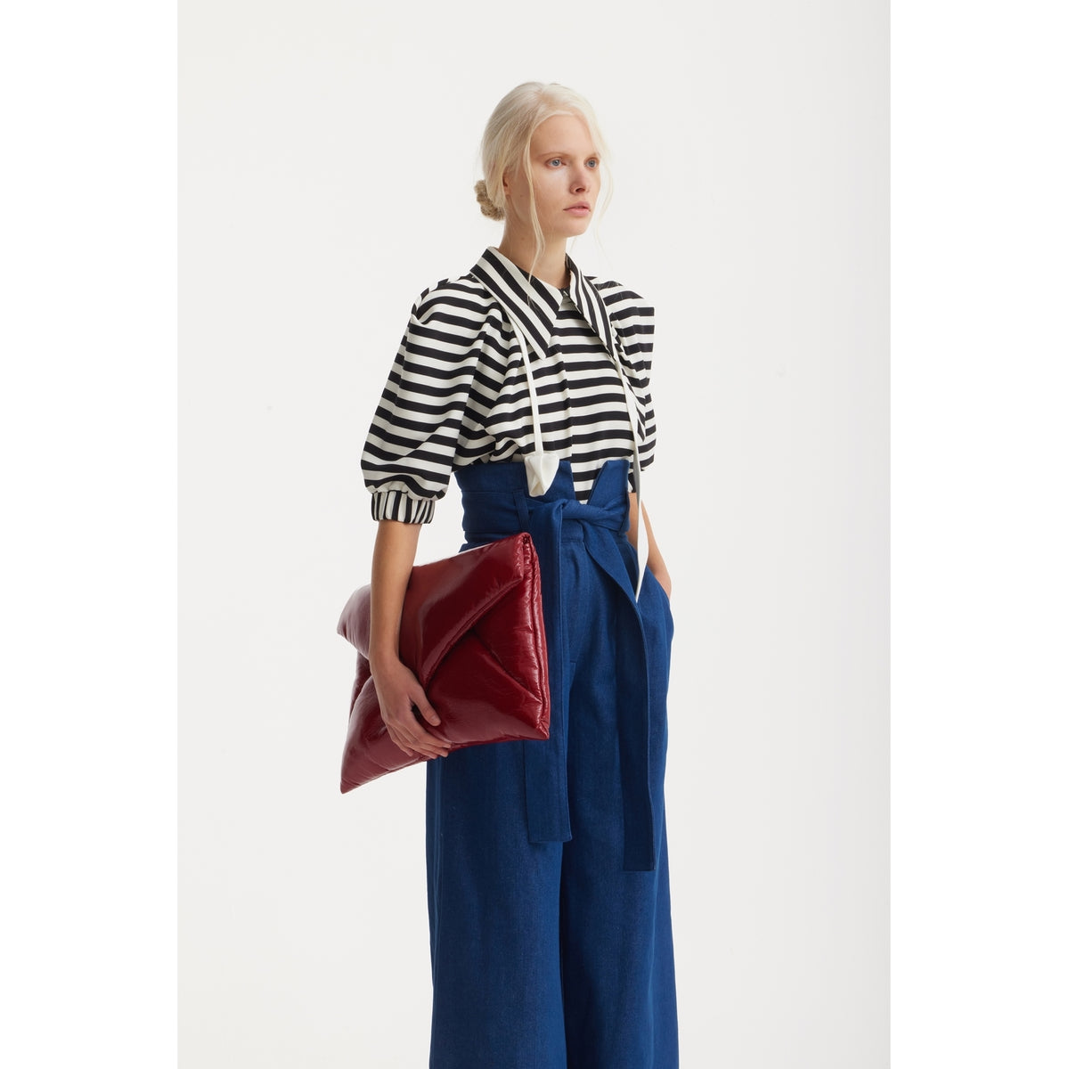 A woman with light blond braided hair wears a JULIA ALLERT Designer Blouse With Black White Stripes and high-waisted blue pants, holding a shiny red clutch bag against a plain white background.