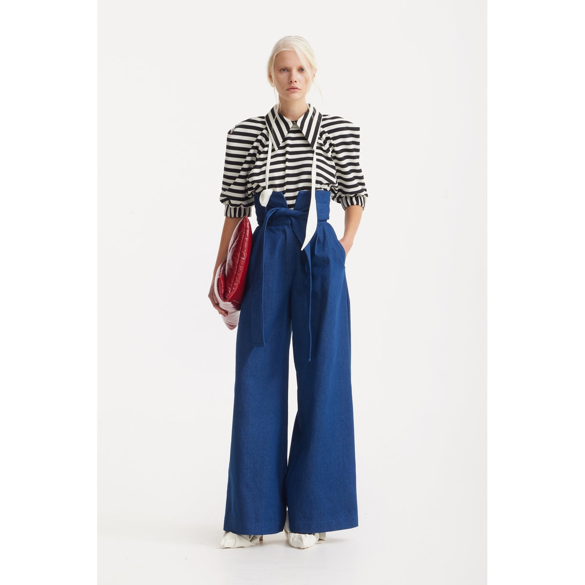 A person faces forward in a JULIA ALLERT Designer Blouse With Black White Stripes, high-waisted wide-leg blue trousers with a bow, white shoes, and holding a shiny red clutch against a plain white background.