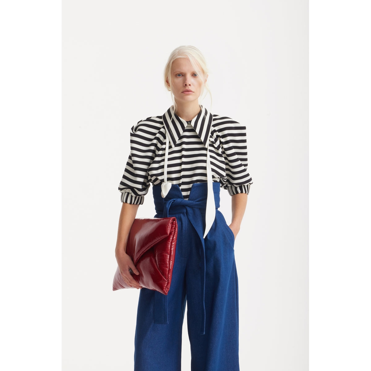 A person with light hair wears a JULIA ALLERT Designer Blouse With Black White Stripes, blue high-waisted pants, and holds a large red clutch against a plain white background.