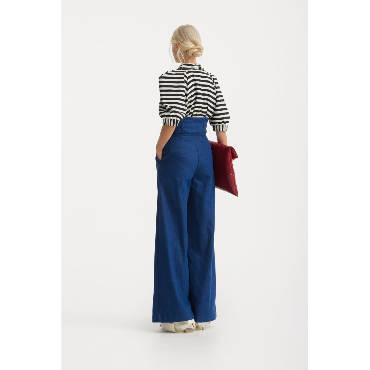 Back view of a woman with her hair in a bun, wearing a JULIA ALLERT Designer Blouse With Black White Stripes, high-waisted blue wide-leg pants, and holding a red clutch.