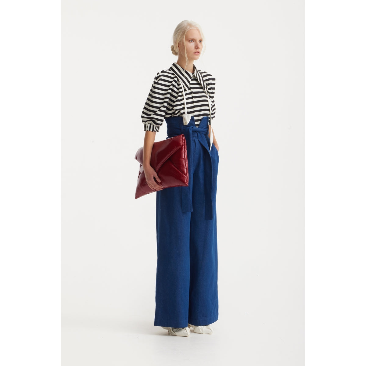 A person stands against a white background wearing a JULIA ALLERT Designer Blouse With Black White Stripes, high-waisted wide-leg blue pants with a tied belt, white shoes, and holding a large red clutch bag.
