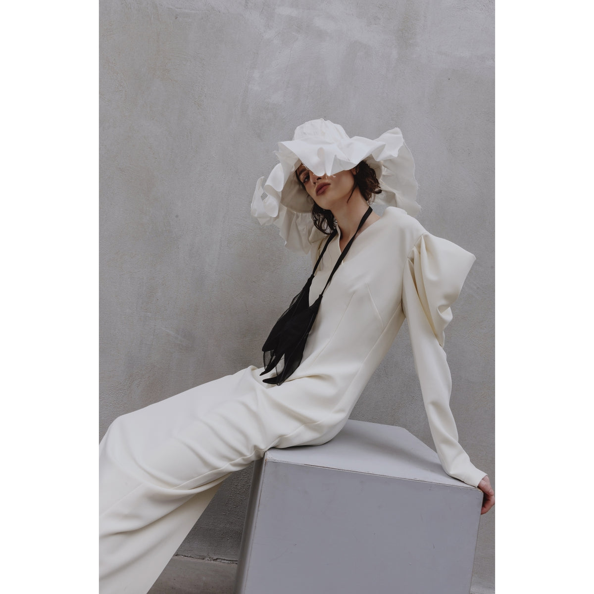 A person in a JULIA ALLERT Crepe Midi Dress With Statement Sleeves - Ivory and a large ruffled white hat with a black ribbon sits on a gray block against a textured light gray wall.