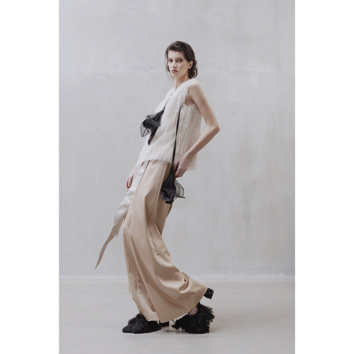 A woman poses against a plain, light wall wearing a white fringe top, JULIA ALLERT High-Waist Wrap Trousers in beige, black feathered slip-ons, and a black sheer waist accent.