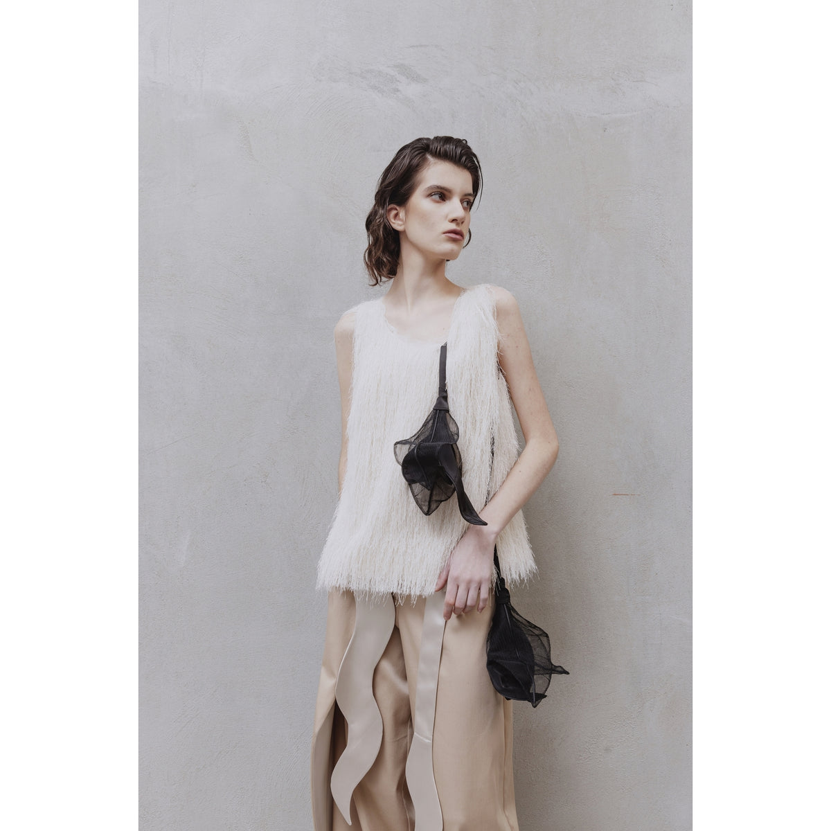 A person with short brown hair wears the JULIA ALLERT Fringe Sleeveless Top in Ivory, paired with beige wide-leg pants and a small black mesh bag, standing before a textured light gray wall.