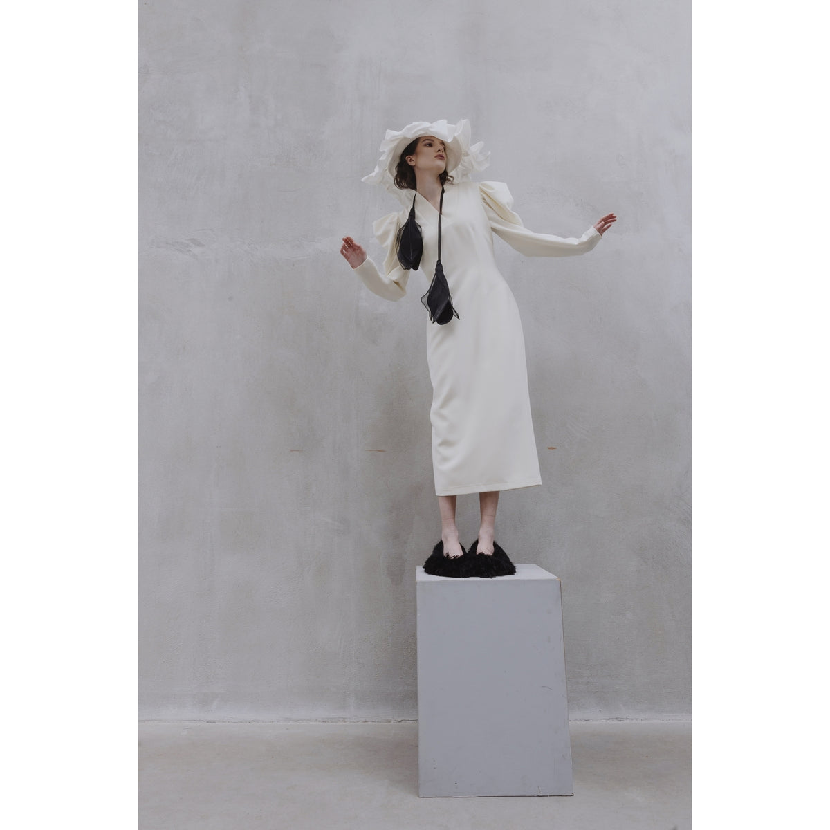 A woman stands on a gray pedestal against a light-gray textured wall, posing with outstretched arms in the JULIA ALLERT Crepe Midi Dress With Statement Sleeves - Ivory, styled with a black tie, wide-brimmed white hat, and black shoes.