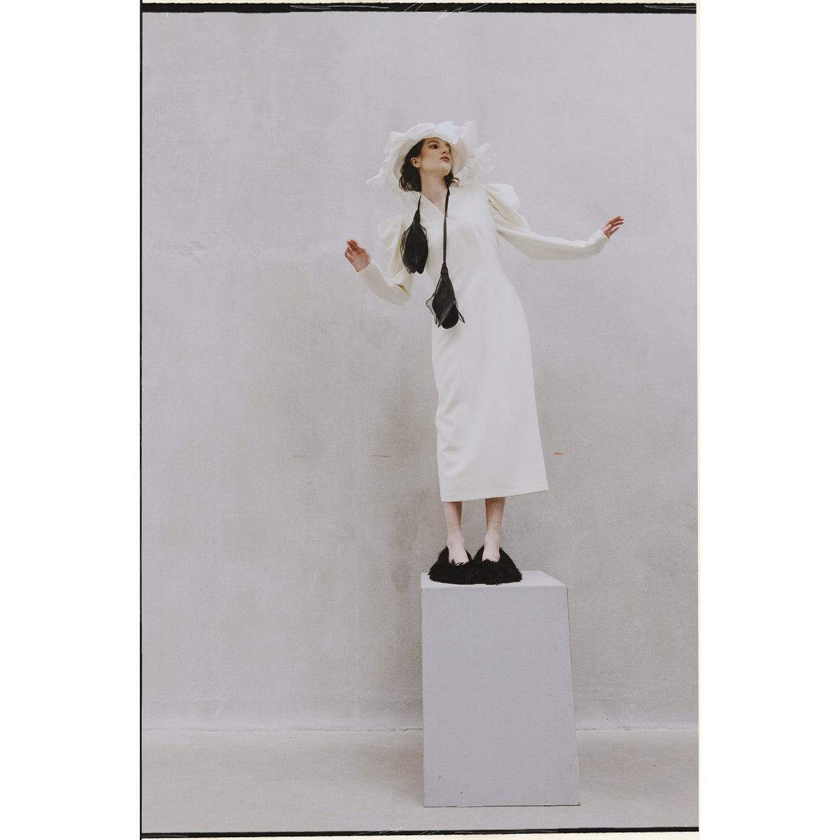A woman wears the JULIA ALLERT Crepe Midi Dress With Statement Sleeves in ivory, styled with a black necktie, large white hat, and black shoes as she poses on a pedestal against a light wall, one arm raised and head tilted back.