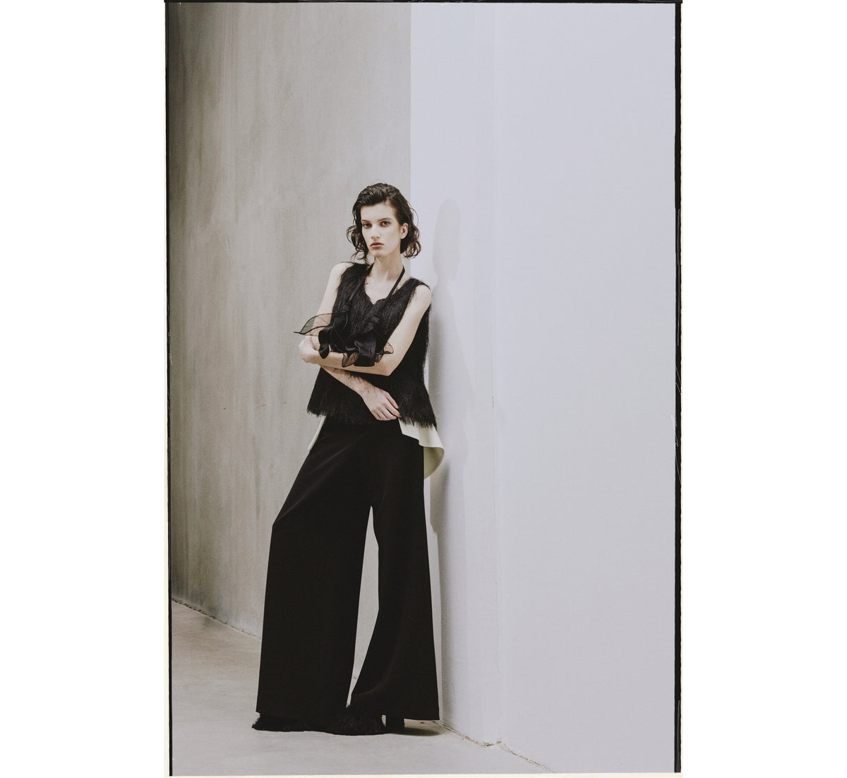 A woman with dark wavy hair stands against a modern, minimalist wall, wearing a JULIA ALLERT Fringe Sleeveless Top - Black and wide-leg black pants. She gazes forward with a neutral expression.