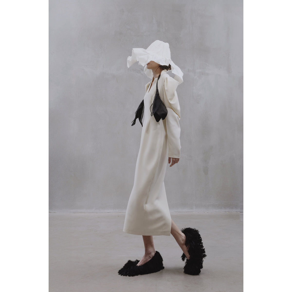 A person wears the JULIA ALLERT Crepe Midi Dress With Statement Sleeves in ivory, paired with sculptural black accessories, oversized textured black slippers, and a large white face-covering headpiece, walking past a plain gray wall.