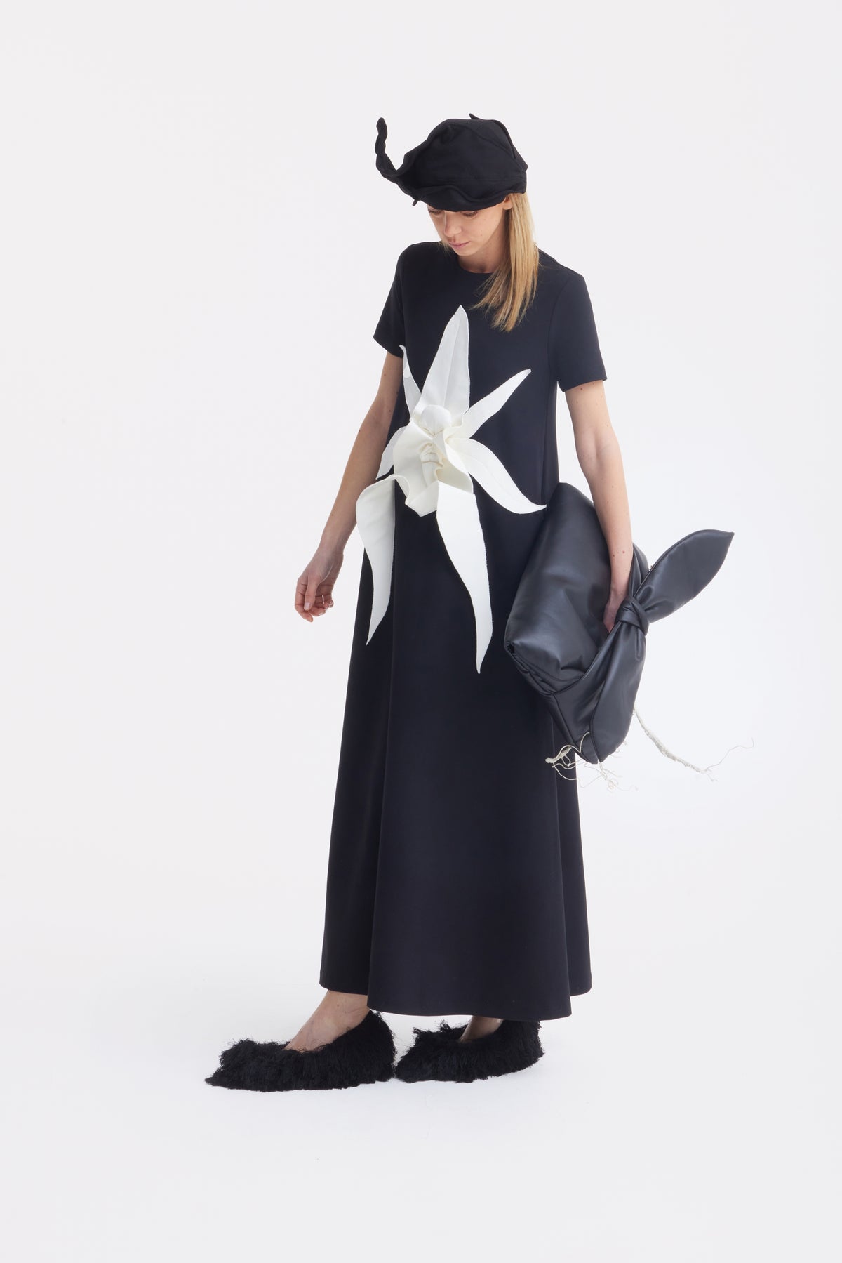 A woman in a long black dress with a white flower applique holds a black cylindrical bag, wearing fluffy black shoes and the JULIA ALLERT Headpiece - Evening Mini Hat Style - Black, looking down against a plain white background.