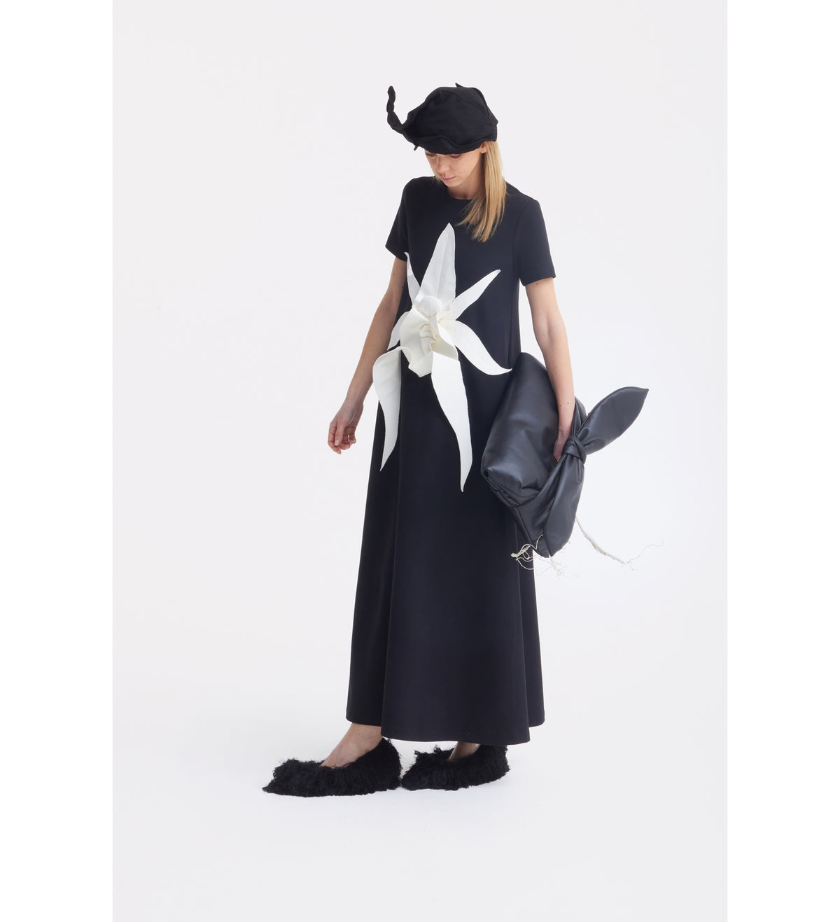 A woman wearing the JULIA ALLERT Ankle-Length Knit Dress With Flower Detail in black poses against a white background. She pairs it with a black hat, black furry shoes, and holds a long-eared black rabbit pillow.