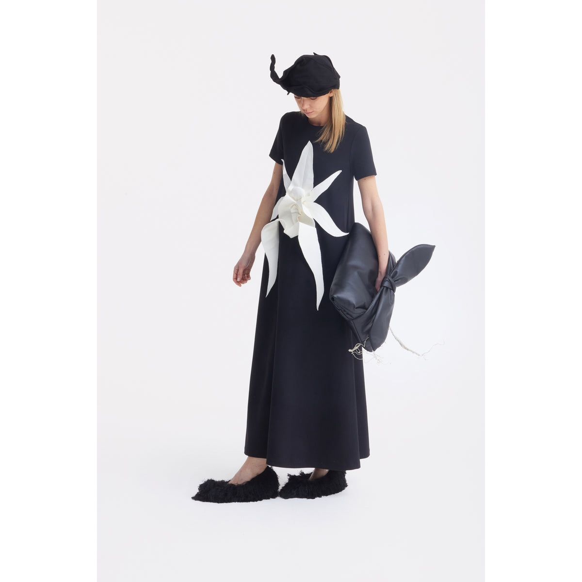 A person in a long black dress with a white flower, black hat, and fuzzy black shoes holds the JULIA ALLERT Petal Faux Leather Evening Clutch Bag - Black against a plain white background.