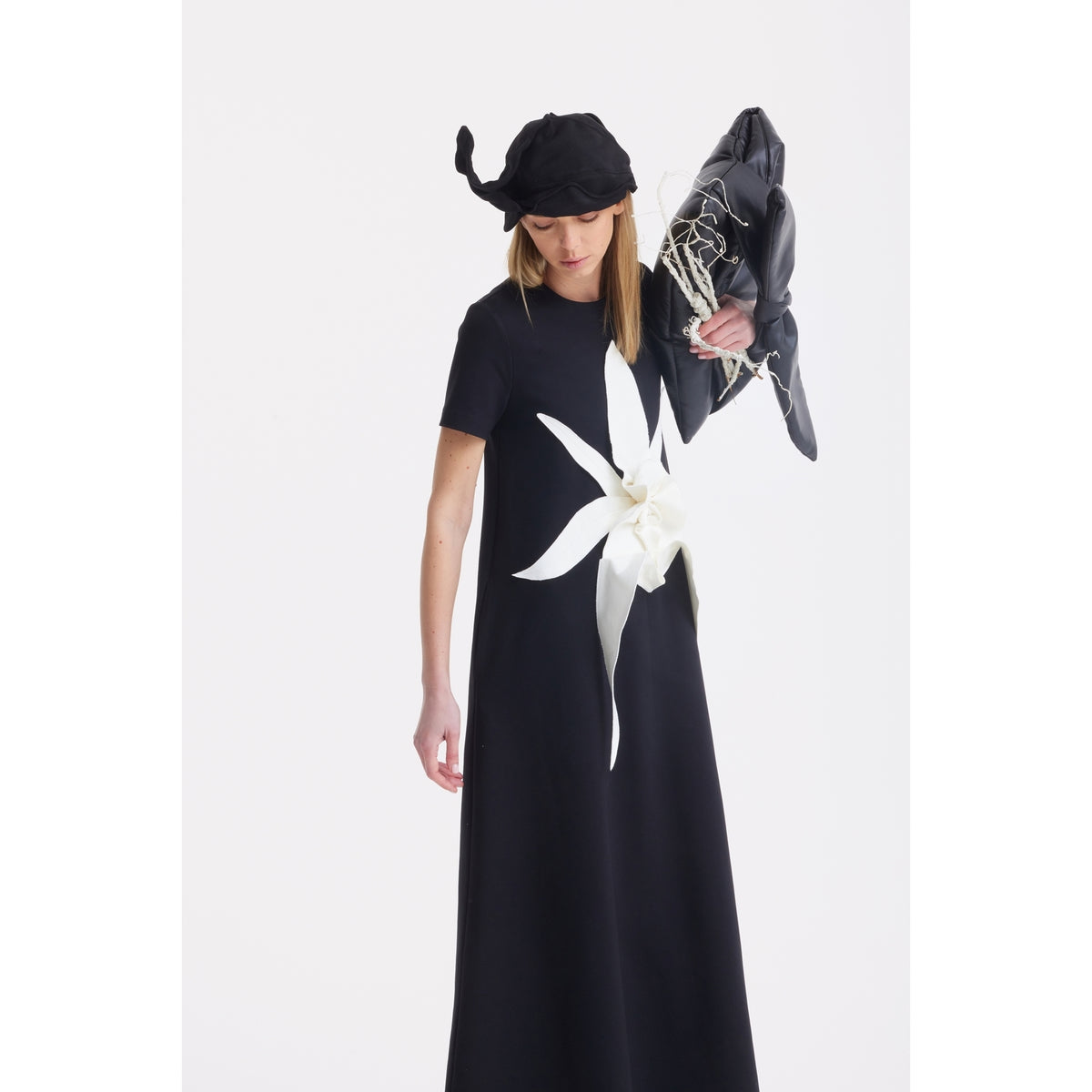 A woman in a long black dress with bold white floral design and a black hat holds the JULIA ALLERT Petal Faux Leather Evening Clutch Bag in black, draped with white cords, standing against a plain white background.