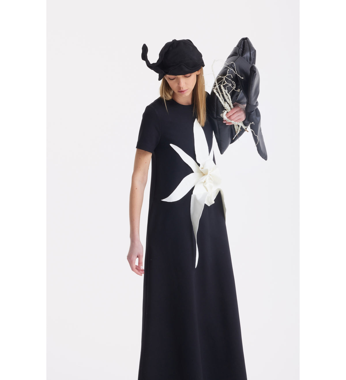 A person wears the JULIA ALLERT Ankle-Length Knit Dress With Flower Detail - Black, a black hat with a pointed curl, and holds a black bag with netted white details against a plain white background.