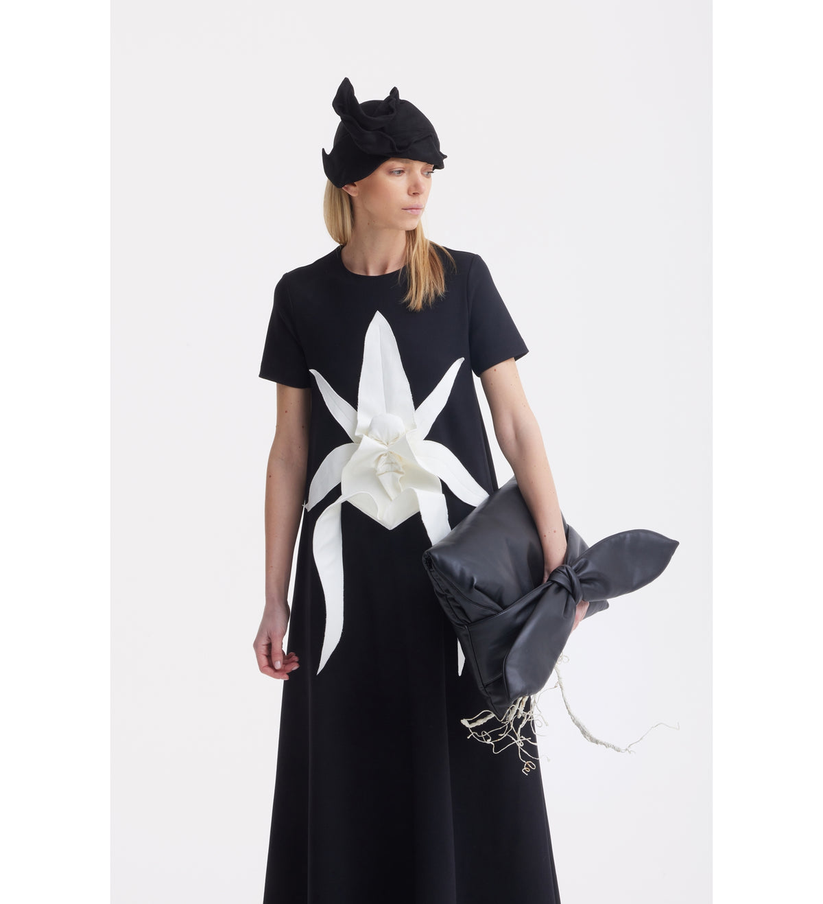 A woman wears the black JULIA ALLERT Ankle-Length Knit Dress With Flower Detail, paired with a black bow-adorned hat and a black clutch with a bow, standing against a plain white background.