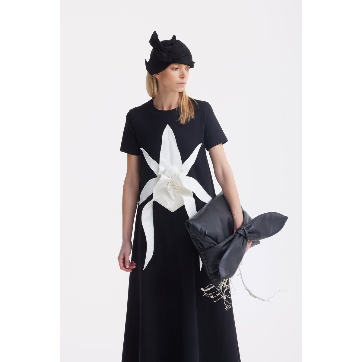 A woman in a black dress with a bold white floral print and pointed black hat holds the JULIA ALLERT Petal Faux Leather Evening Clutch Bag - Black, standing against a plain white background.