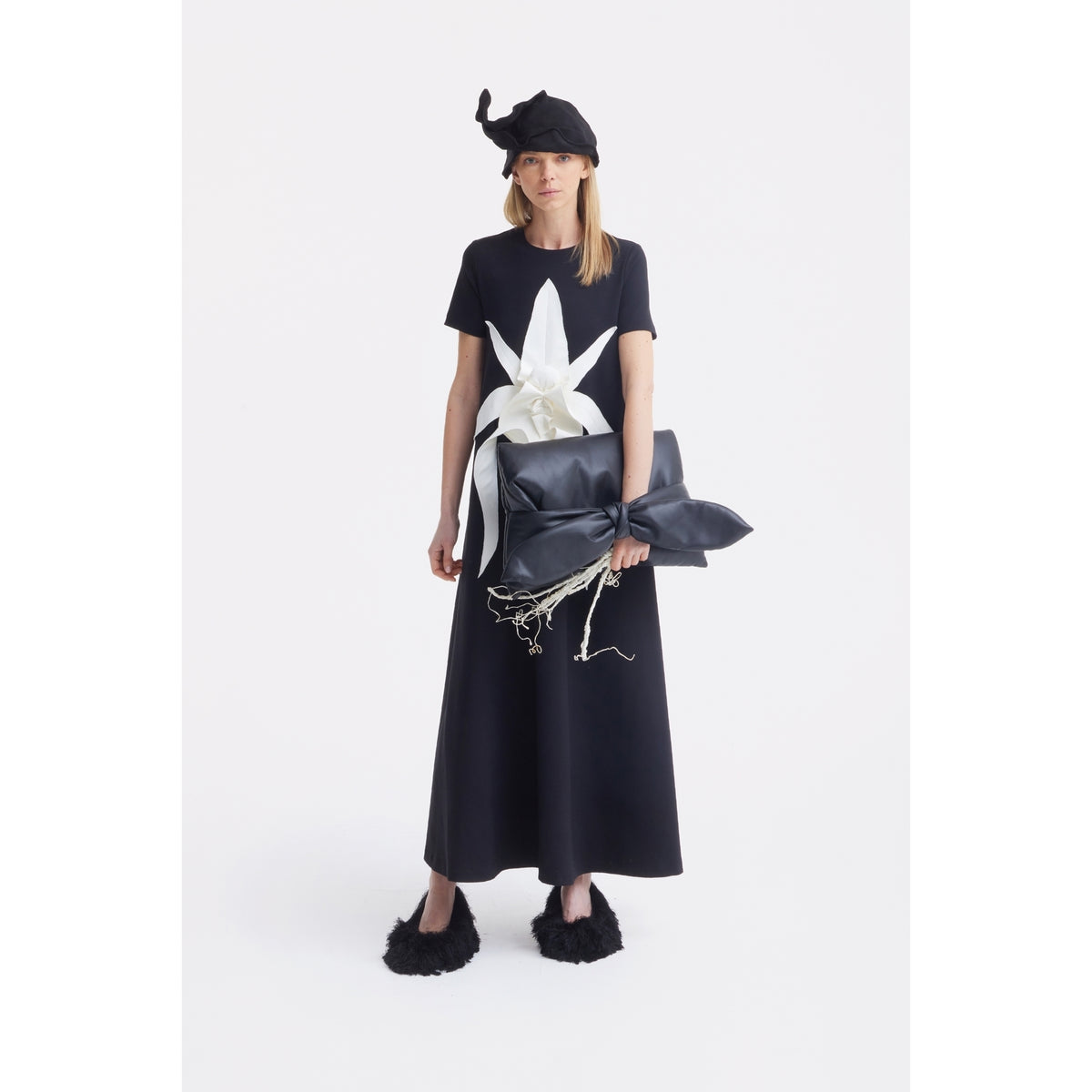 A person in a long black dress with a large white bow holds an oversized black clutch with a white ribbon and wears the JULIA ALLERT Headpiece - Evening Mini Hat Style - Black, standing against a plain white background.
