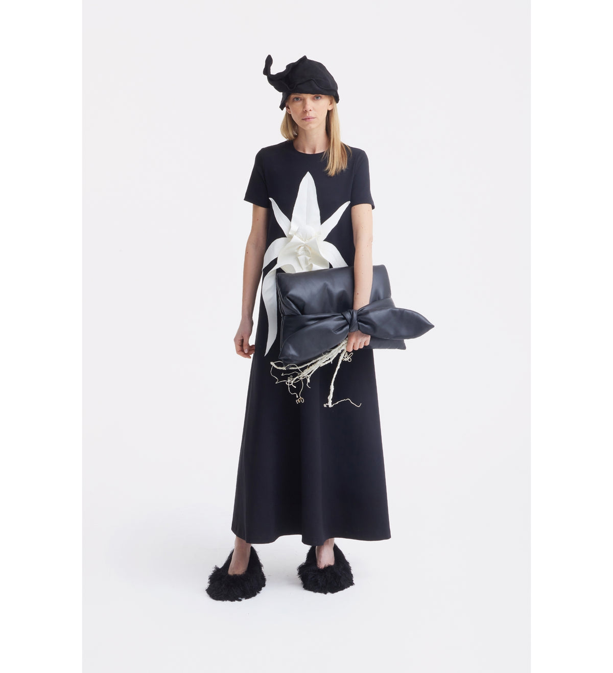 A woman in the JULIA ALLERT Ankle-Length Knit Dress With Flower Detail - Black stands against a white background, styled with a black hat, fuzzy shoes, a black bow bag, and dried white branches.