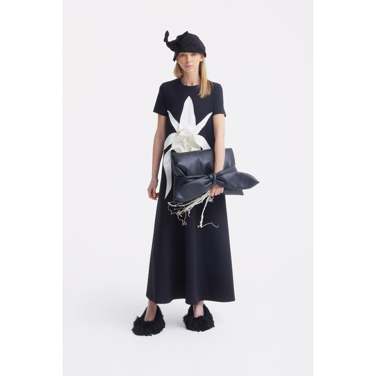 A woman in a long black dress with a white bow at the waist holds the JULIA ALLERT Petal Faux Leather Evening Clutch Bag - Black. She wears a pointed black hat and fuzzy shoes, standing against a plain white background.
