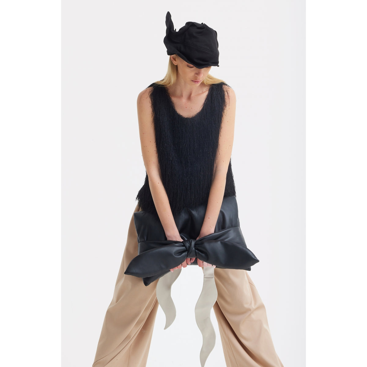 A person wears a black textured sleeveless top, JULIA ALLERT High-Waist Wrap Trousers in beige, and a sculptural black hat, holding a large black bow with long white ribbon tails against a plain white background.