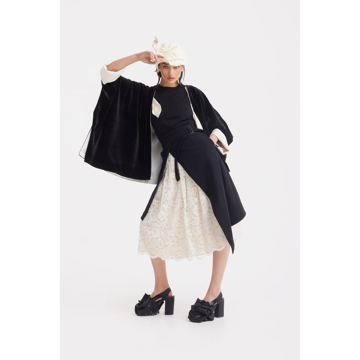 A woman poses in a chic black and white outfit featuring the JULIA ALLERT Loose Velvet Cape Jacket With Zip Front, a lace skirt, black belt, chunky heels, and a unique white hat against a plain white background.