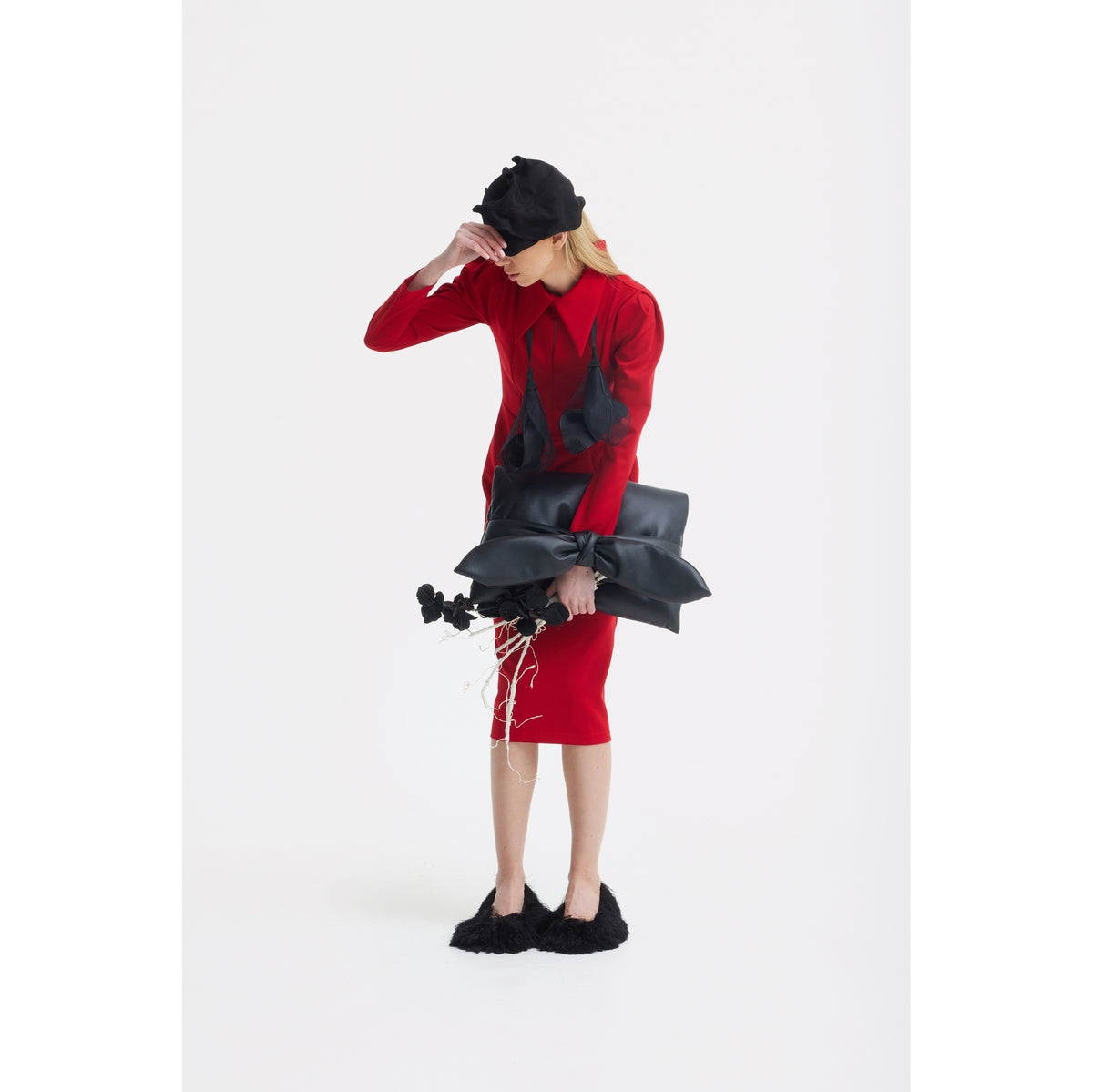 Wearing a JULIA ALLERT Solid Formal Midi Dress in red and a black hat, a person stands against a white background holding black sculptural objects and dried flowers. They wear black furry shoes and partially cover their face with one hand.