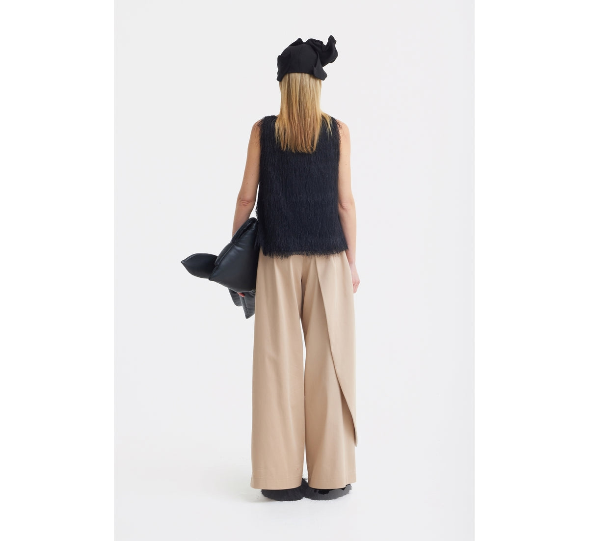 A woman faces away, wearing the JULIA ALLERT Fringe Sleeveless Top in black, loose beige wide-leg pants, a black headscarf, and holding a black bag against a plain white background.