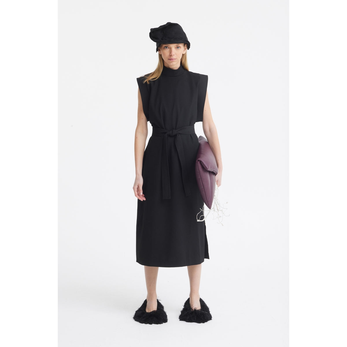 A woman stands against a white background wearing a sleeveless black dress with a belt, furry black slippers, and the JULIA ALLERT Headpiece - Evening Mini Hat Style in black. She carries a large purple clutch bag, perfect as an evening accessory.