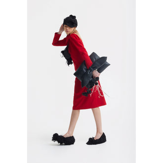 A woman in a JULIA ALLERT Solid Formal Midi Dress - Red with a black hat poses against a white background, holding a large abstract black accessory. She wears black furry shoes and looks over her shoulder, one hand touching her hat.