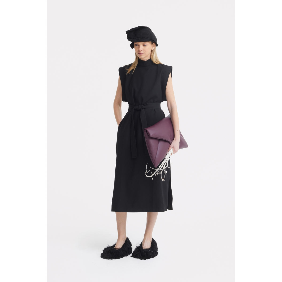 A woman poses against a white background in a sleeveless black dress, black fluffy slippers, and the JULIA ALLERT Headpiece - Evening Mini Hat Style in Black, finishing her look with a large purple clutch adorned with a white accent.