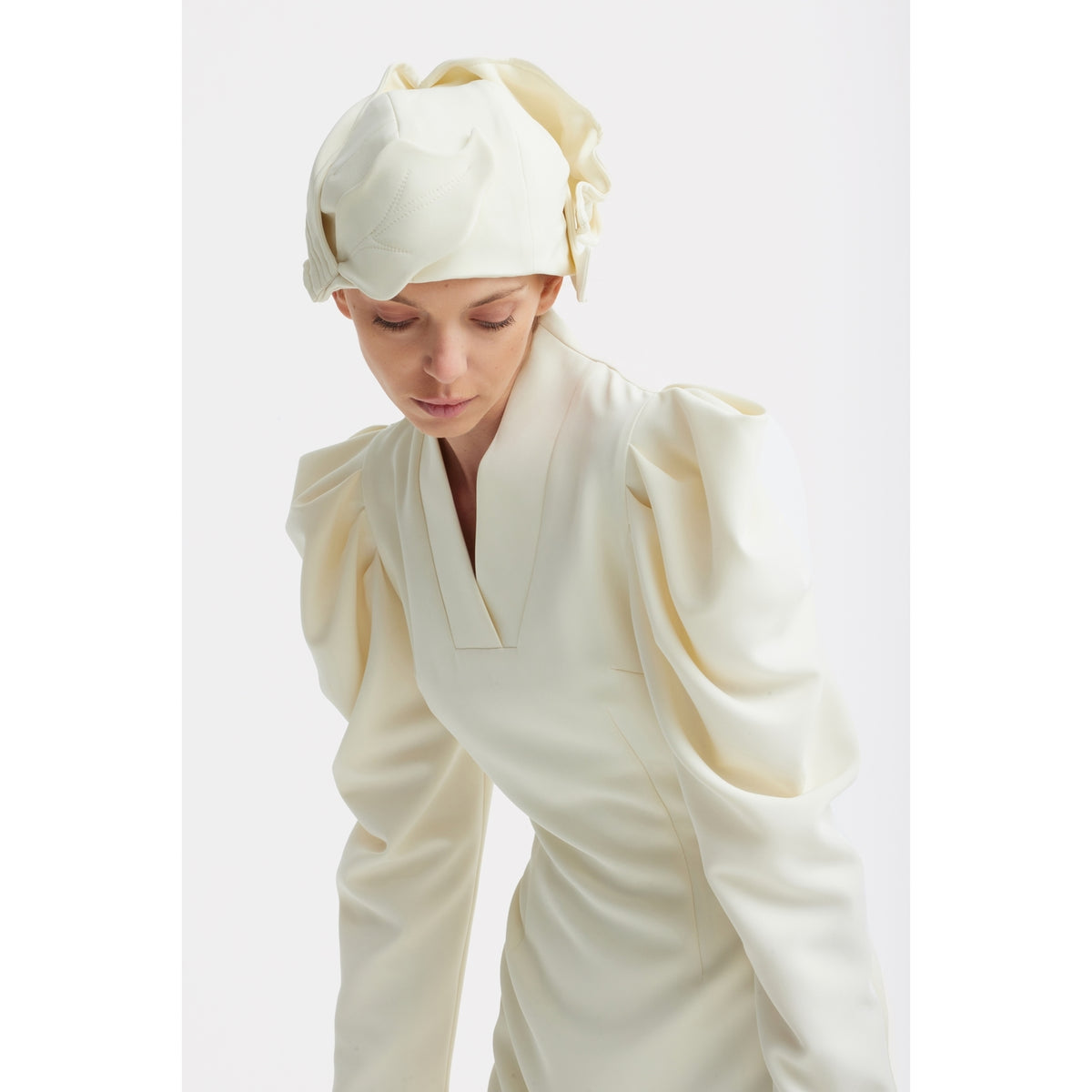 A woman in the JULIA ALLERT Crepe Midi Dress With Statement Sleeves - Ivory and a matching sculpted headpiece looks down against a plain white background.