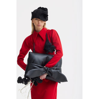 A woman in a JULIA ALLERT Solid Formal Midi Dress - Red and black hat holds a large black handbag with an oversized bow. She has long blonde hair, a serious expression, and stands against a plain white background.