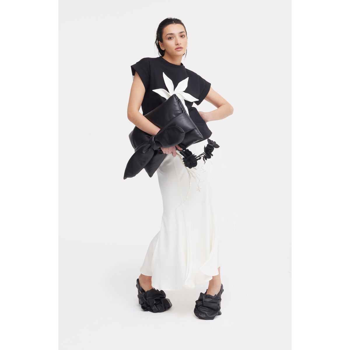 A woman models the JULIA ALLERT Crew-Neck Jersey T-Shirt With Flower in black, styled with a flowing white skirt, black chunky bow shoes, and an oversized black bag against a plain white background.