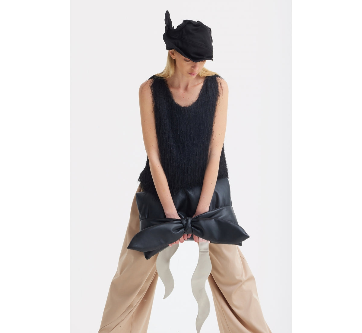 A woman wearing the JULIA ALLERT Fringe Sleeveless Top in black, wide beige pants, and a black abstract hat, holding a large black bow with white ribbon tails, stands against a plain white background.