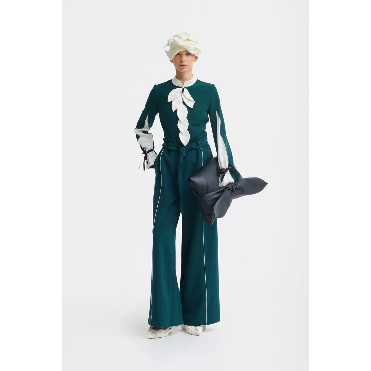 A person stands against a white background in a teal JULIA ALLERT suit with wide-leg trousers, a matching top with white ruffle accents, a sculptural white hat, white shoes, and holds the Petal Faux Leather Evening Clutch Bag - Black by JULIA ALLERT.