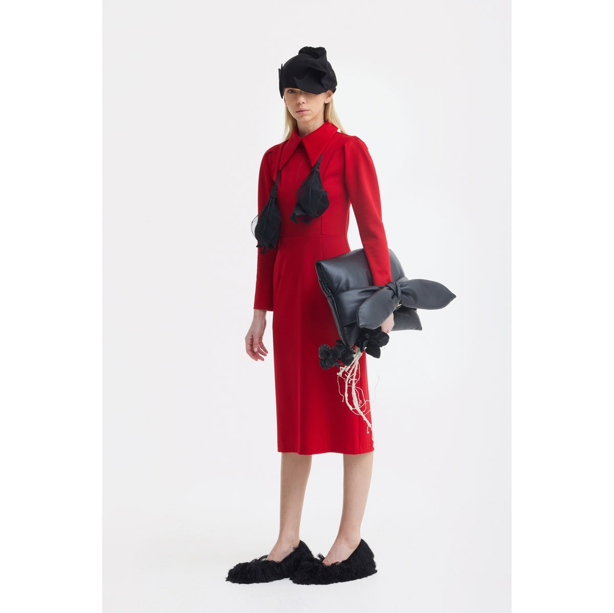 A woman in a JULIA ALLERT Solid Formal Midi Dress - Red, black furry slippers, a black headpiece, and a sculptural black bow-accented bag stands against a plain white background.