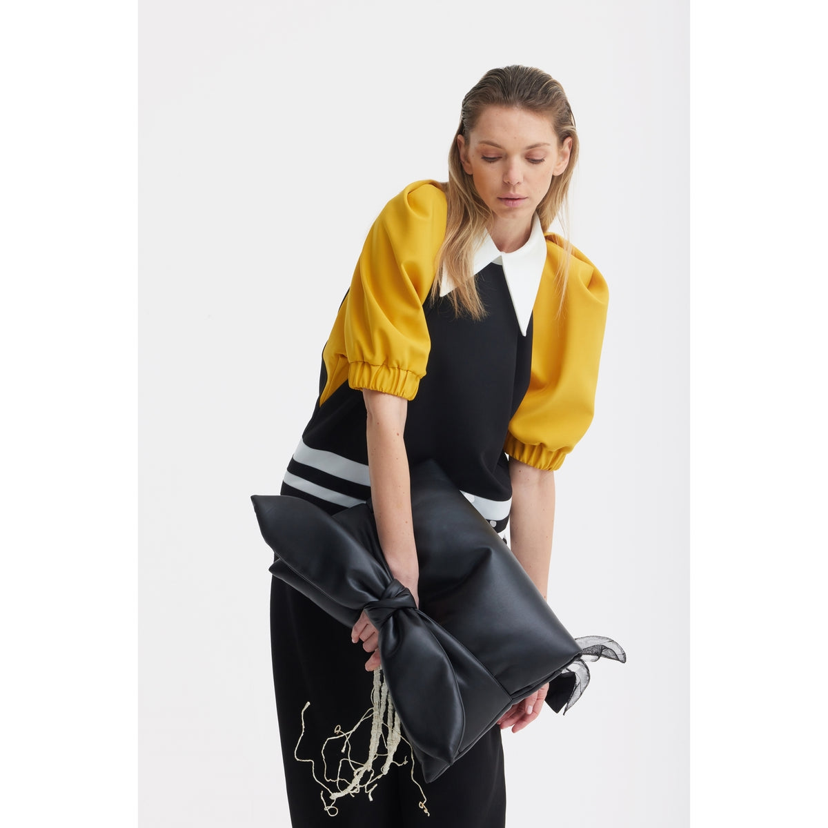 A woman with long blonde hair wears a JULIA ALLERT black top with yellow puff sleeves and a white collar, holding a JULIA ALLERT Petal Faux Leather Evening Clutch Bag - Black shaped like a bow, looking down against a plain white background.