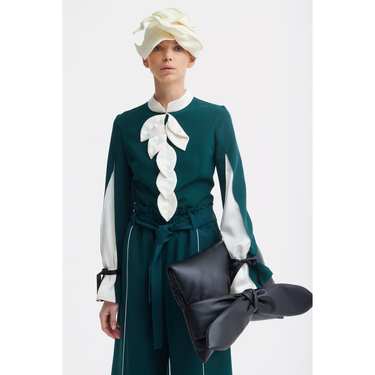 A person in a dark green outfit with white leaf accents and a cream sculptural hat holds the JULIA ALLERT Petal Faux Leather Evening Clutch Bag in black against a plain white background.