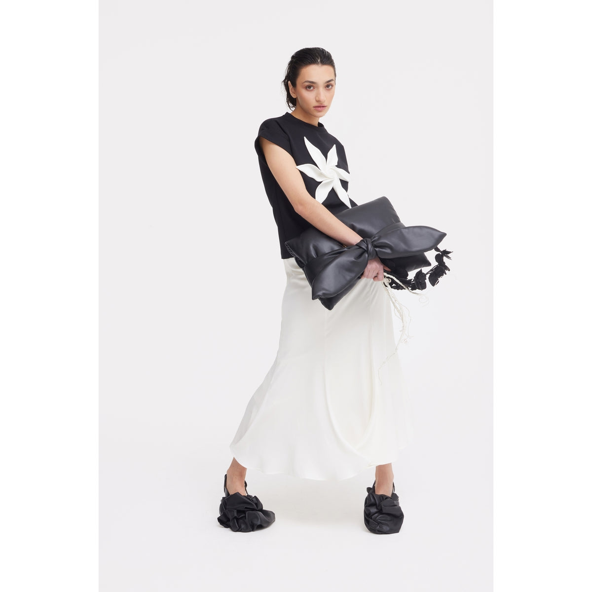 A woman poses against a white background, wearing the JULIA ALLERT Crew-Neck Jersey T-Shirt With Flower in black, a long white skirt, black ruffled shoes, and holding an oversized black bow-shaped clutch.