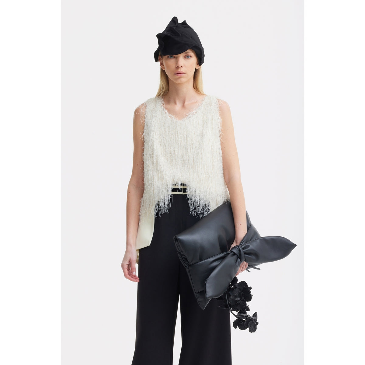 A woman in a textured white sleeveless top, black wide-leg pants, and a black asymmetrical hat holds the JULIA ALLERT Petal Faux Leather Evening Clutch Bag - Black with bow and floral accents against a plain white background.