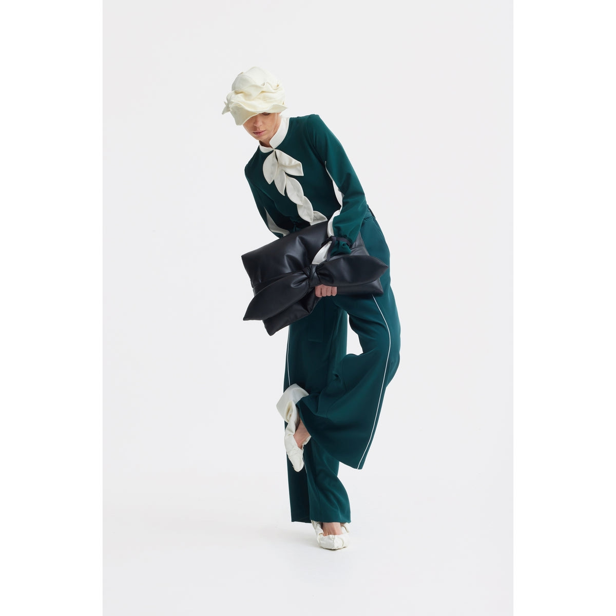 A person in a teal suit with white accents and a large white bow poses holding the JULIA ALLERT Petal Faux Leather Evening Clutch Bag - Black. They wear a floral-detailed white hat and white heels against a plain white background.