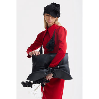 A woman in a JULIA ALLERT Solid Formal Midi Dress - Red and black hat holds a large black bow bag and carries black artificial flowers. The background is plain white.