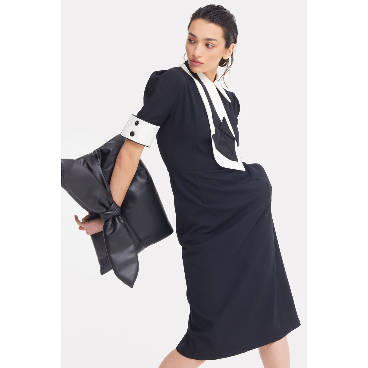 A woman in a black JULIA ALLERT midi dress with puffed sleeves and a geometric black-and-white front poses holding the Petal Faux Leather Evening Clutch Bag in black by JULIA ALLERT, featuring a stylish bow detail.