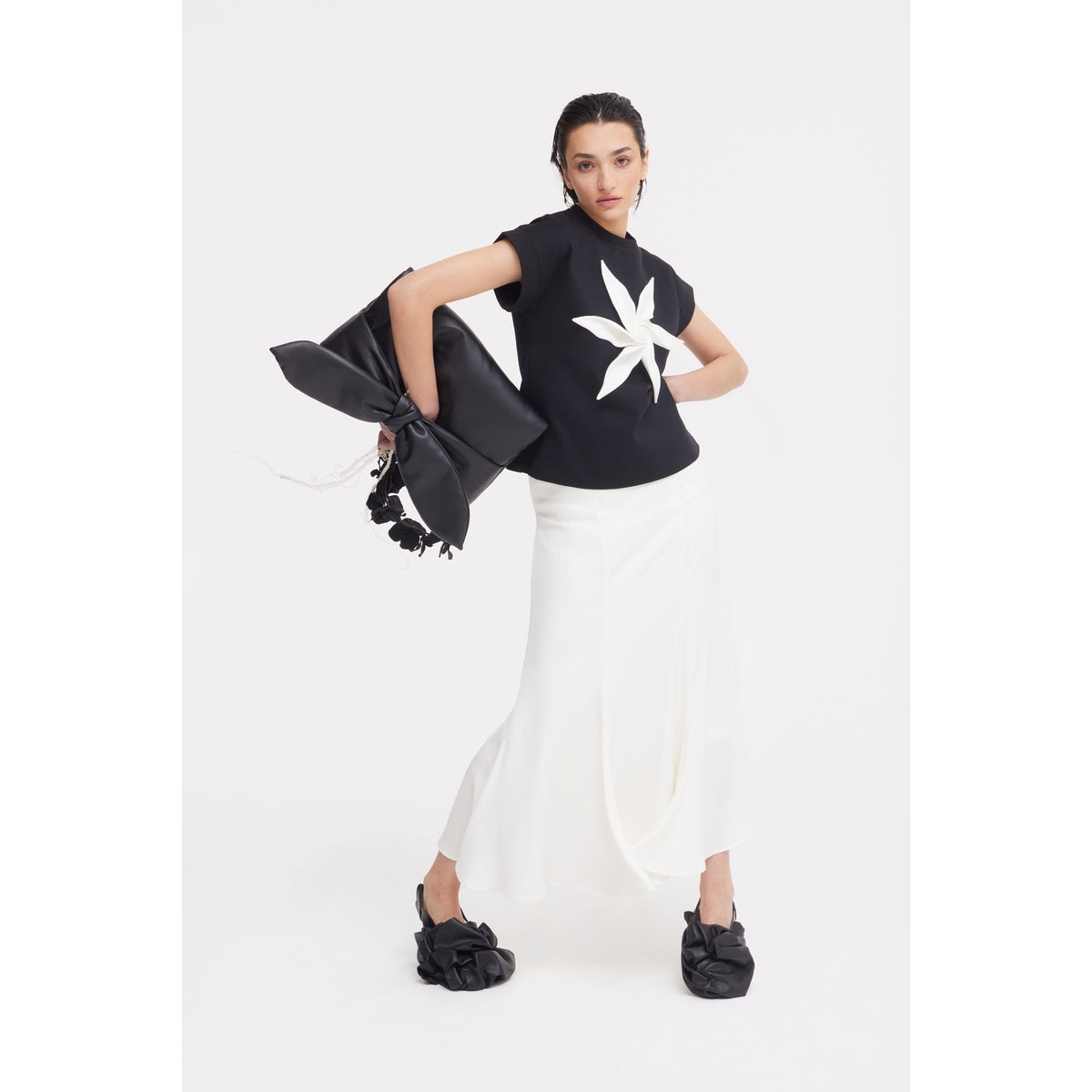 A woman poses in the JULIA ALLERT Crew-Neck Jersey T-Shirt With Flower - Black, styled with a flowing white skirt, black bow-detailed shoes, and a large black bag decorated with bows and flowers, against a plain white background.