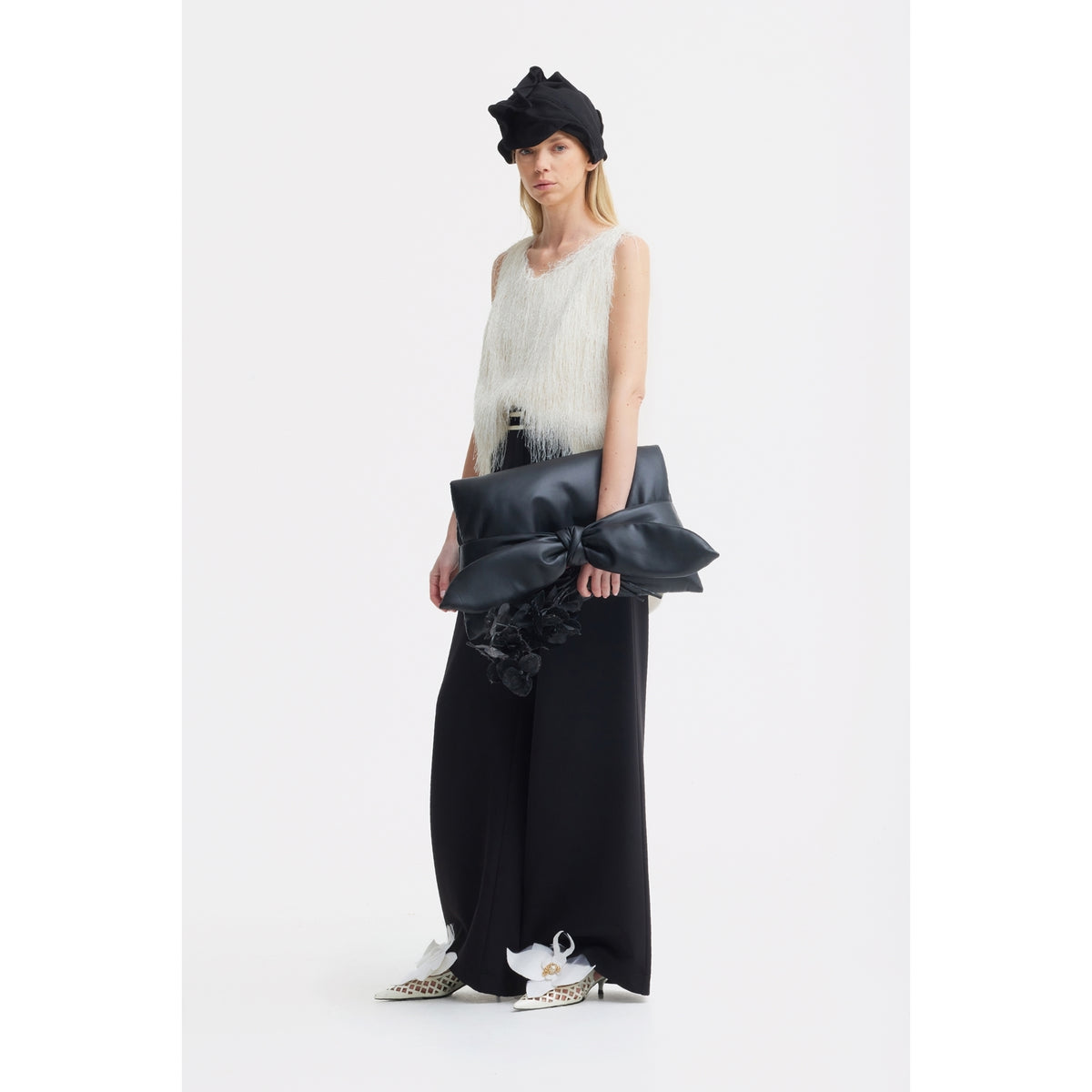 A woman in a white sleeveless textured top, wide black pants, white pointed-toe shoes, and a black headwrap stands against a white background holding the JULIA ALLERT Petal Faux Leather Evening Clutch Bag - Black with a big bow.